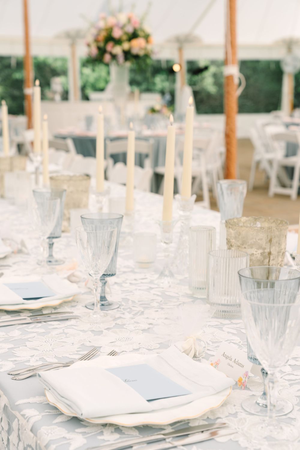 Elegant wedding reception table set with crystal glasses, taper candles, white linens, and floral centerpieces in a tent venue.