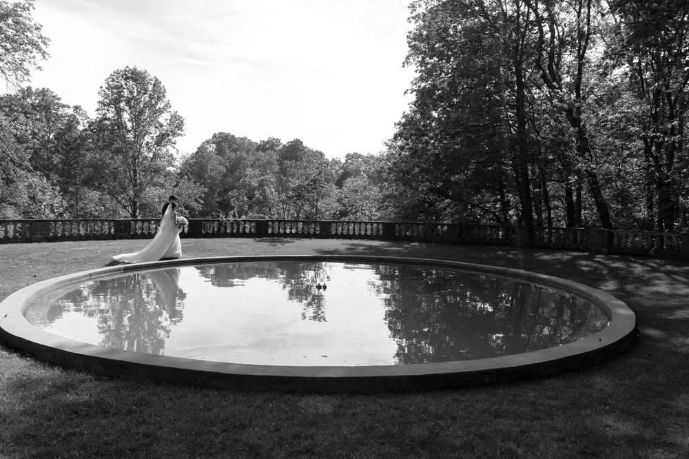Black and white photo of a bride standing beside a circular reflecting pool surrounded by trees at an elegant outdoor venue.