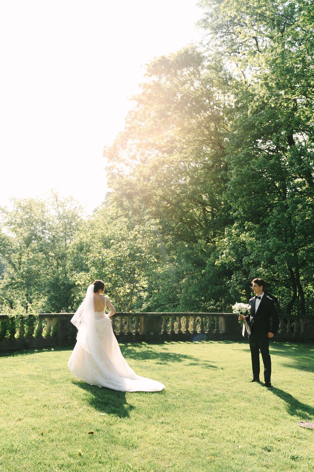 Bride and groom share a romantic first look on a sunlit green lawn surrounded by lush trees at an outdoor wedding.