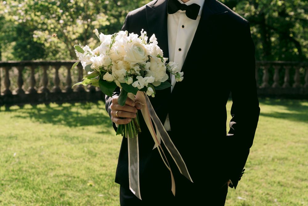 Groom in black tuxedo holding elegant white wedding bouquet with ribbon streamers on green lawn.
