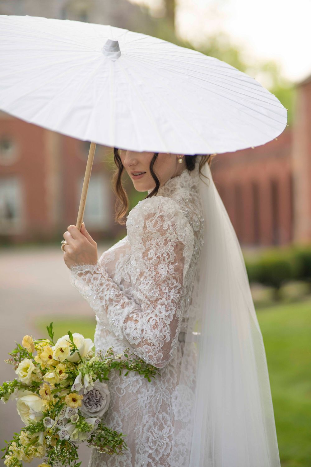 Bride in delicate lace wedding dress holds white parasol umbrella and yellow floral bouquet in garden setting.