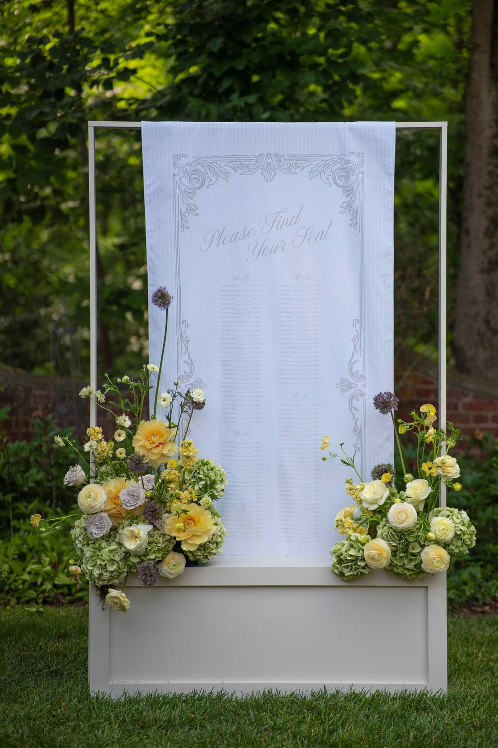 Elegant wedding seating chart display with yellow and white floral arrangements on a white board in a garden setting.