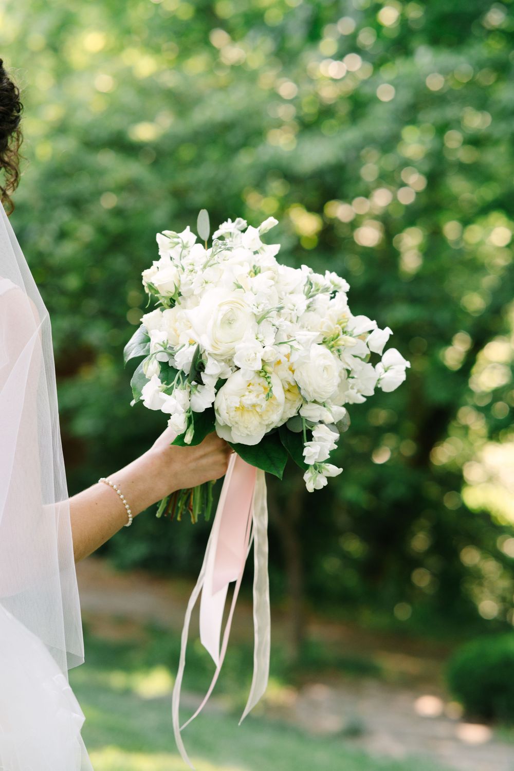 Bride holding a lush white peony and ranunculus wedding bouquet with pink satin ribbons outdoors.