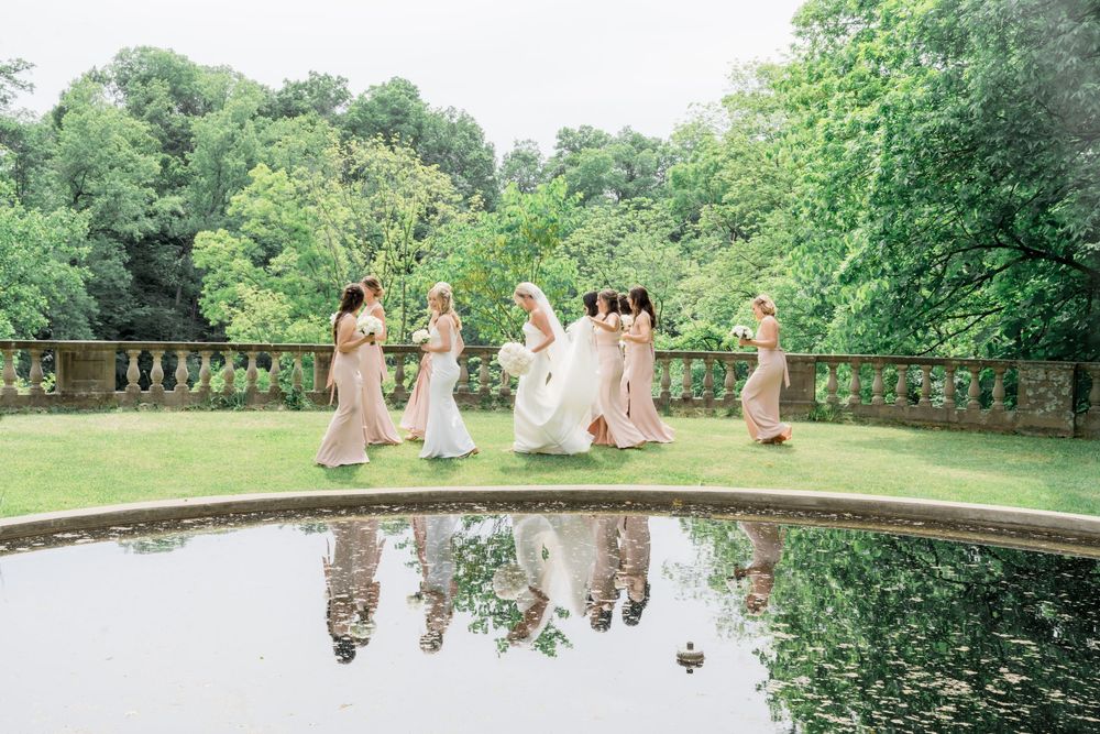 A group in flowing dresses dances beside a reflective pond on a lush garden terrace with stone balustrades.