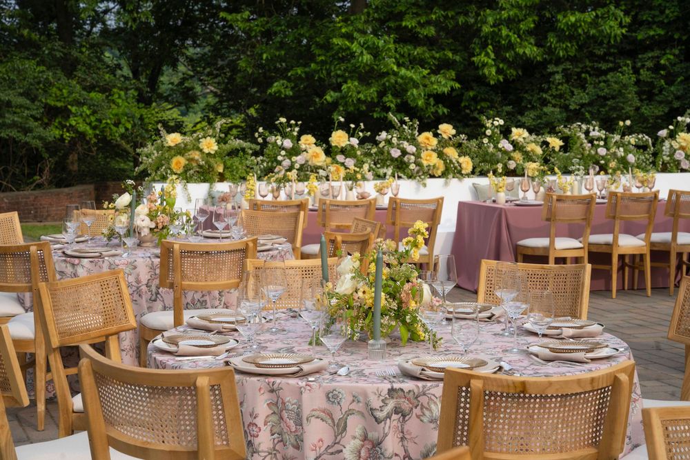 Elegant outdoor wedding reception table setting with woven chairs and yellow floral centerpieces on patterned linens.