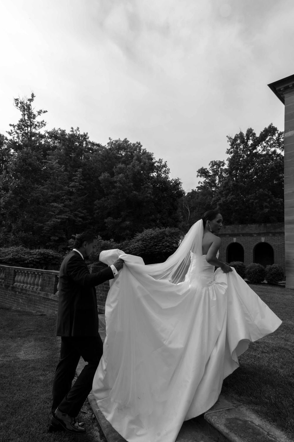 A wedding couple poses outdoors with flowing white dress veil caught in the wind in black and white photography.