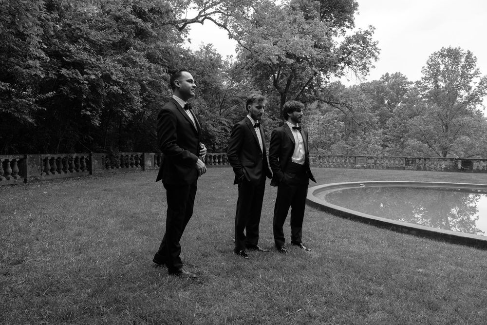 Three men in black tuxedos stand on a lawn near a fountain in a black and white outdoor photo.