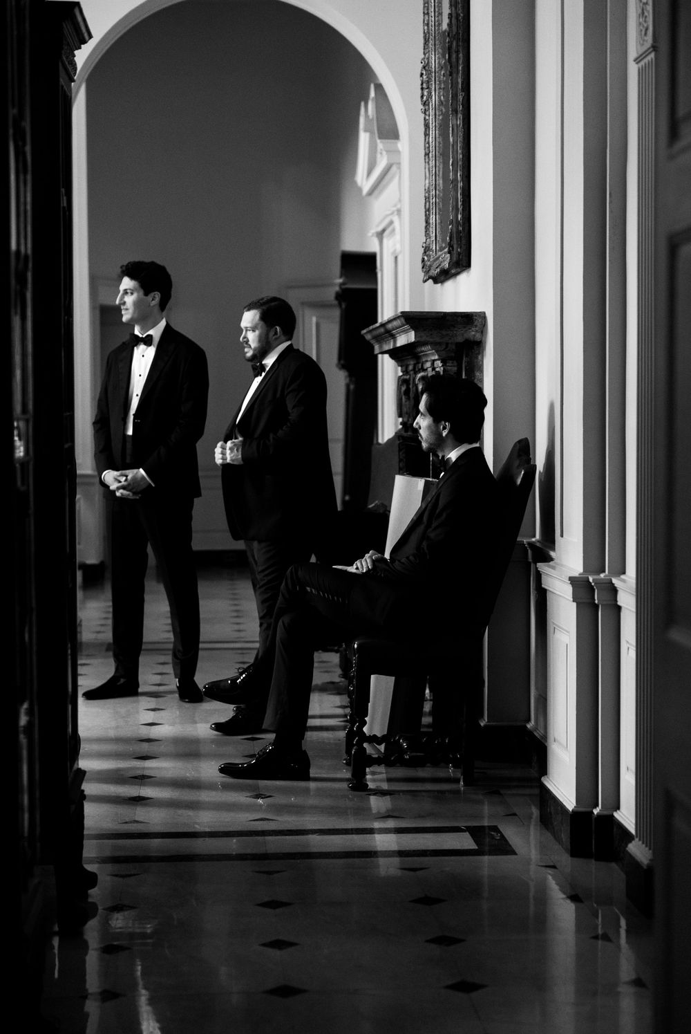 Three groomsmen in black tuxedos waiting in an elegant hallway, captured in dramatic black and white photography.