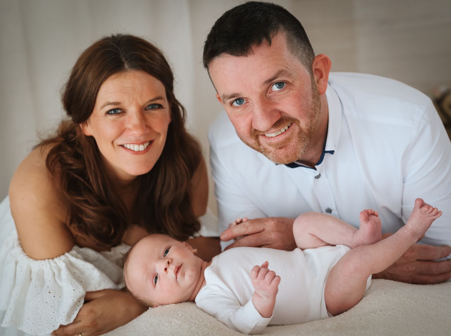Family portrait with baby in white outfit against light background.