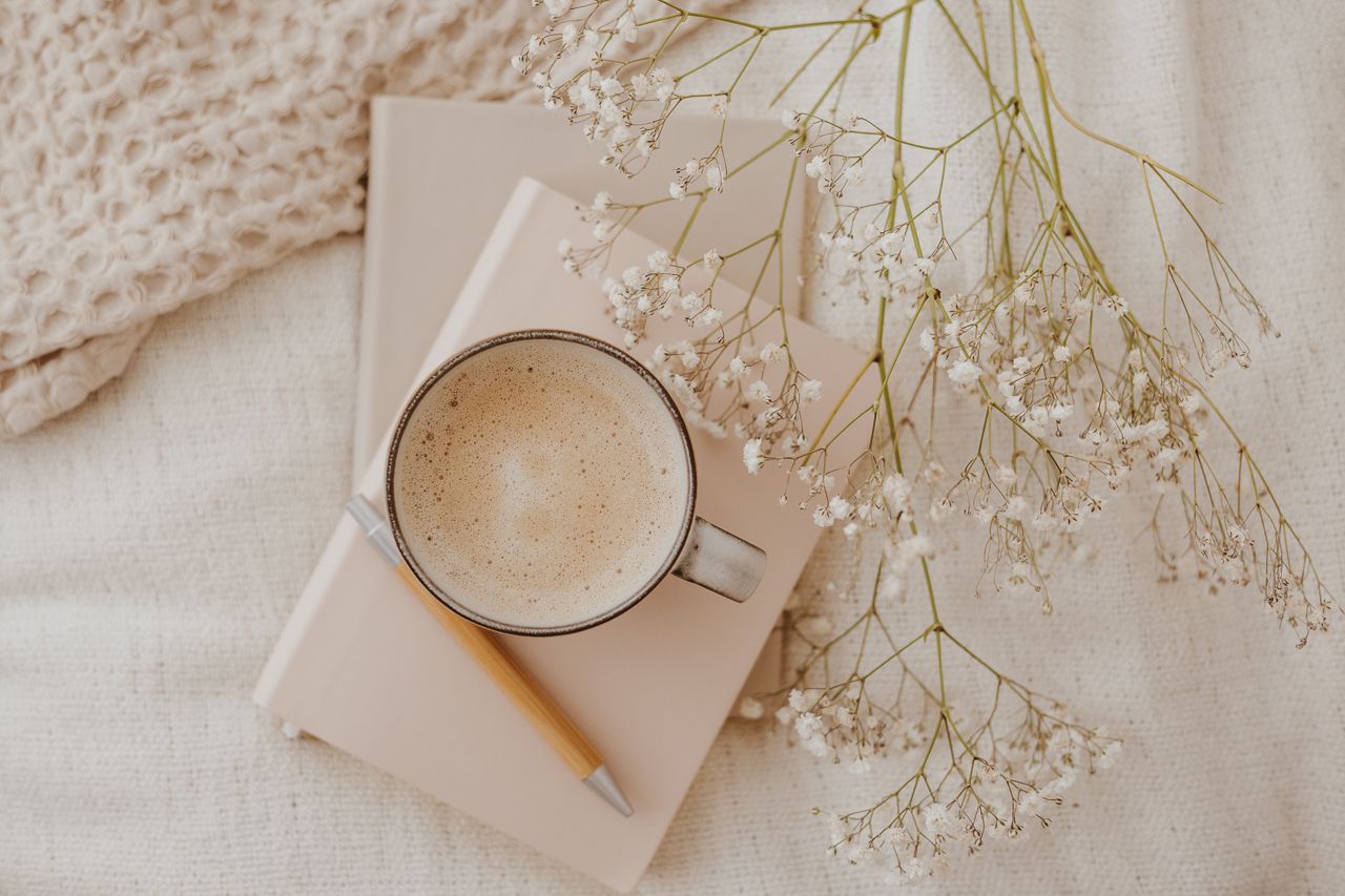 Beige ceramic coffee cup with frothy latte sitting on book with delicate baby's breath flowers on white textile background.