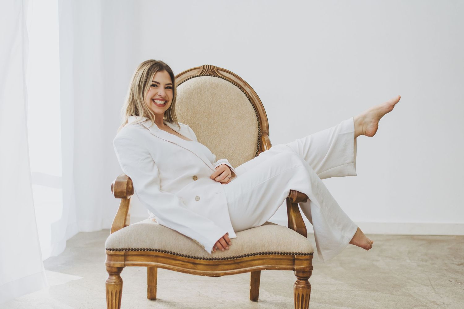 Relaxed woman in white pant suit lounges sideways on vintage beige velvet chair against light neutral background.