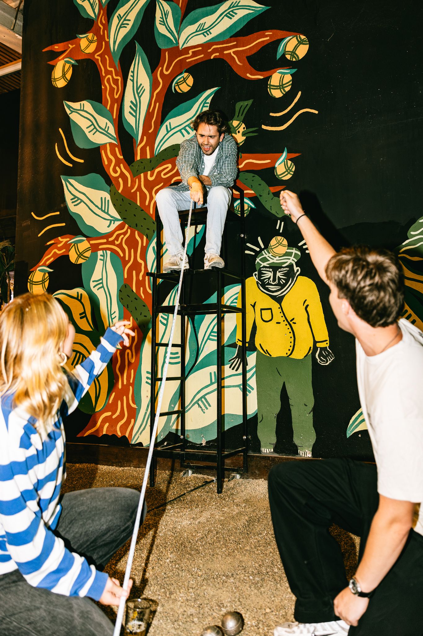 Man perched on ladder in front of colorful tree mural while two people play mini golf below.