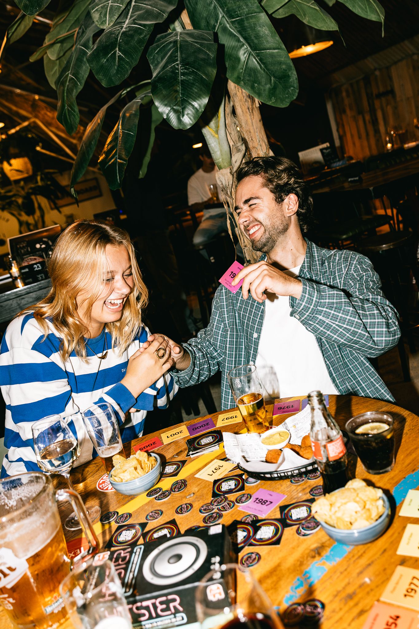 A smiling couple enjoying drinks and snacks at a rustic bar table with a board game and tropical plants nearby.