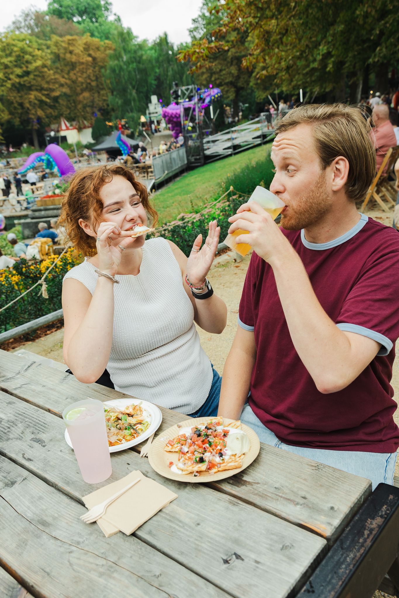 Couple enjoying pizza and beer at an outdoor wooden table near a park canal on a sunny day.