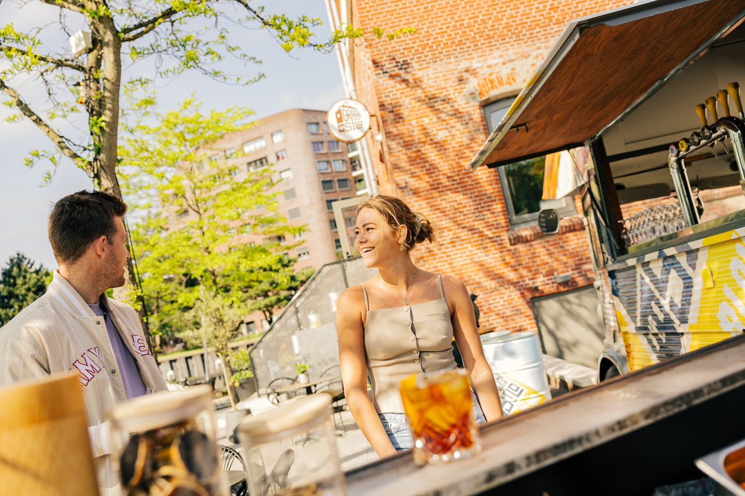 Two people enjoying drinks at an outdoor bar patio on a sunny afternoon with brick walls and trees in the background.