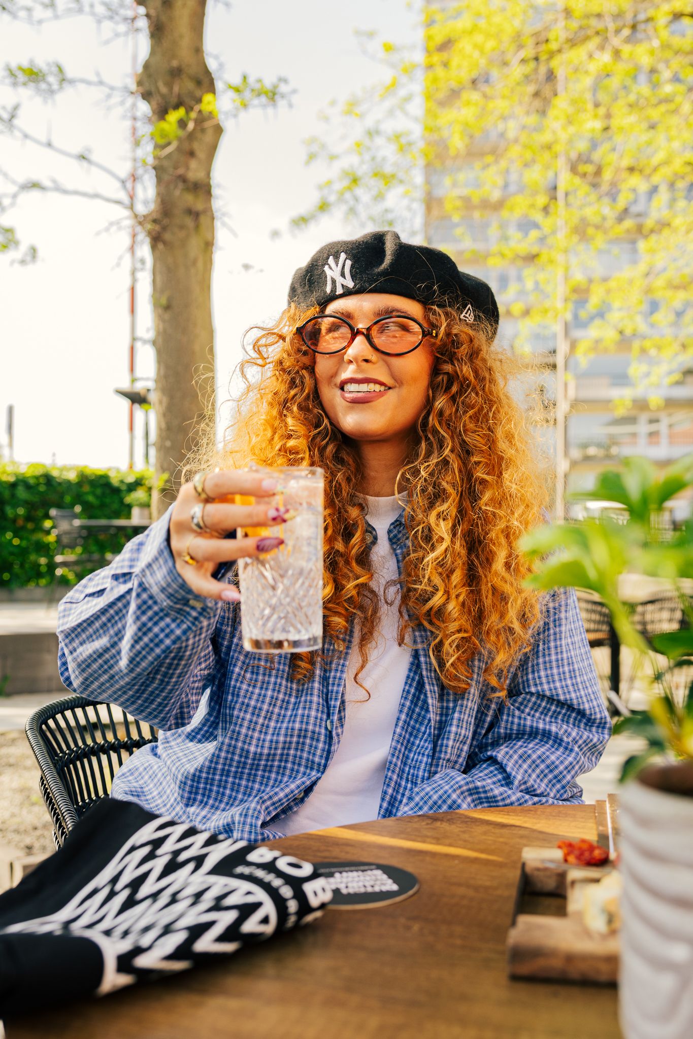Person with curly hair wearing glasses and NY Yankees cap enjoying a cocktail at outdoor restaurant table.