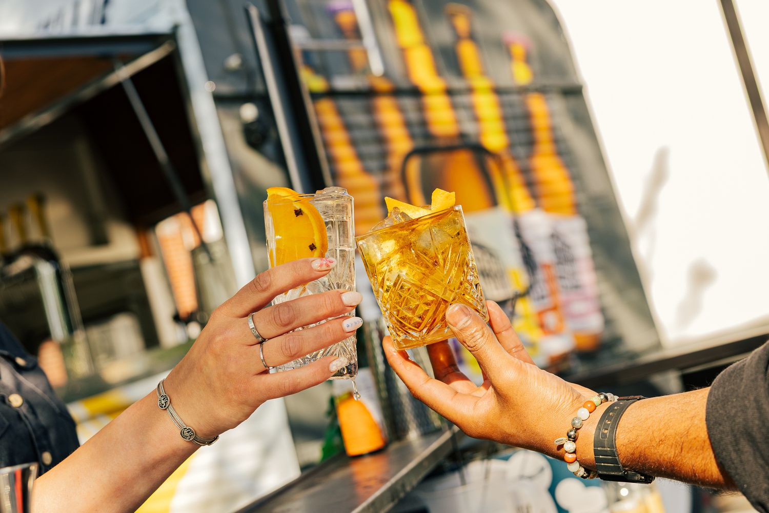 Two hands toasting drinks through a food truck window while holding golden-colored beverages in plastic cups.