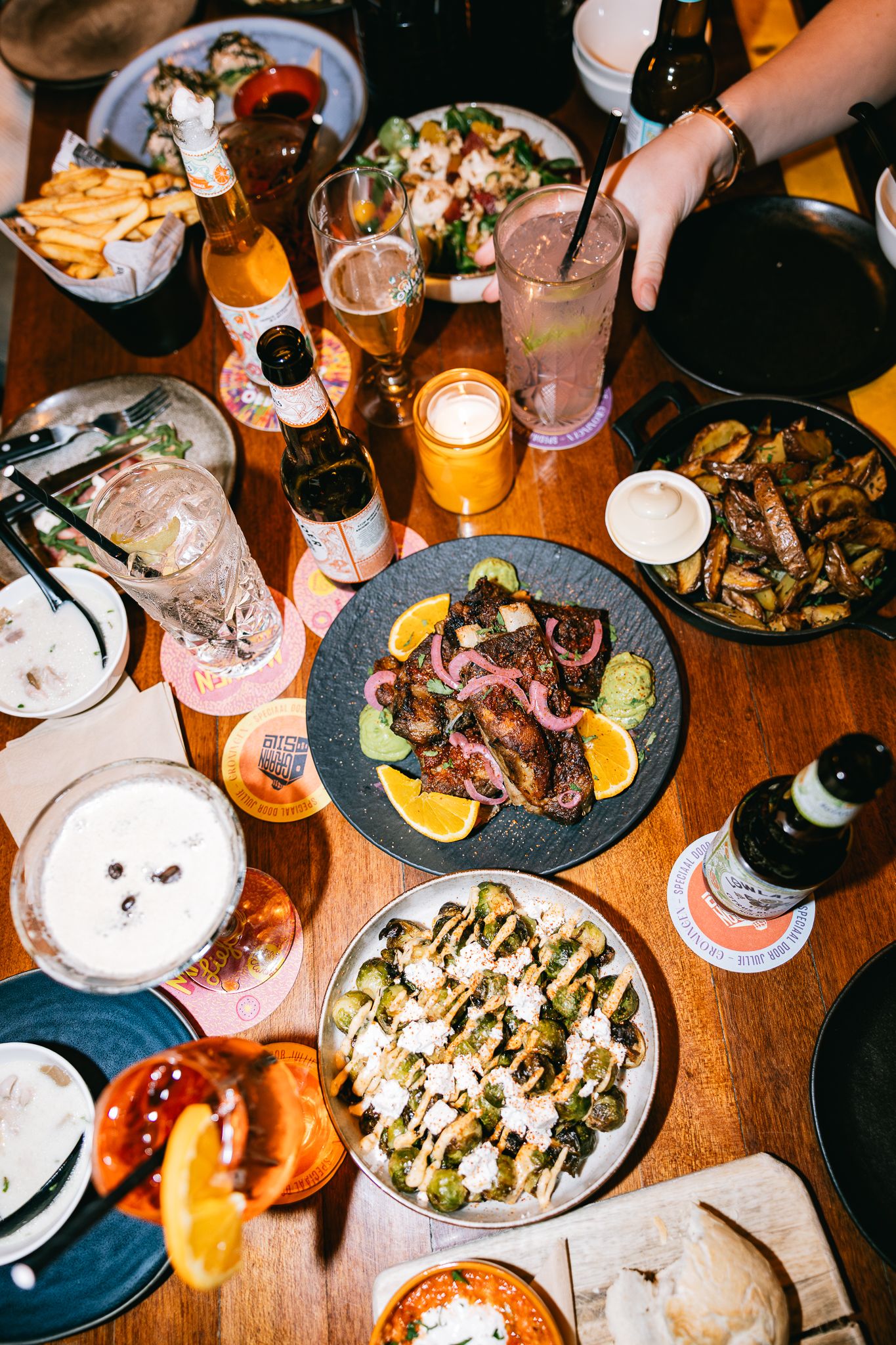 Overhead view of a shared dinner table with plates of food, beer bottles, and cocktails at a restaurant.