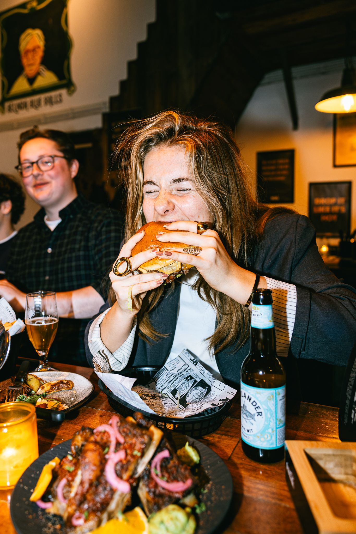 Woman joyfully biting into a large burger at a restaurant table with beer bottle and food nearby.