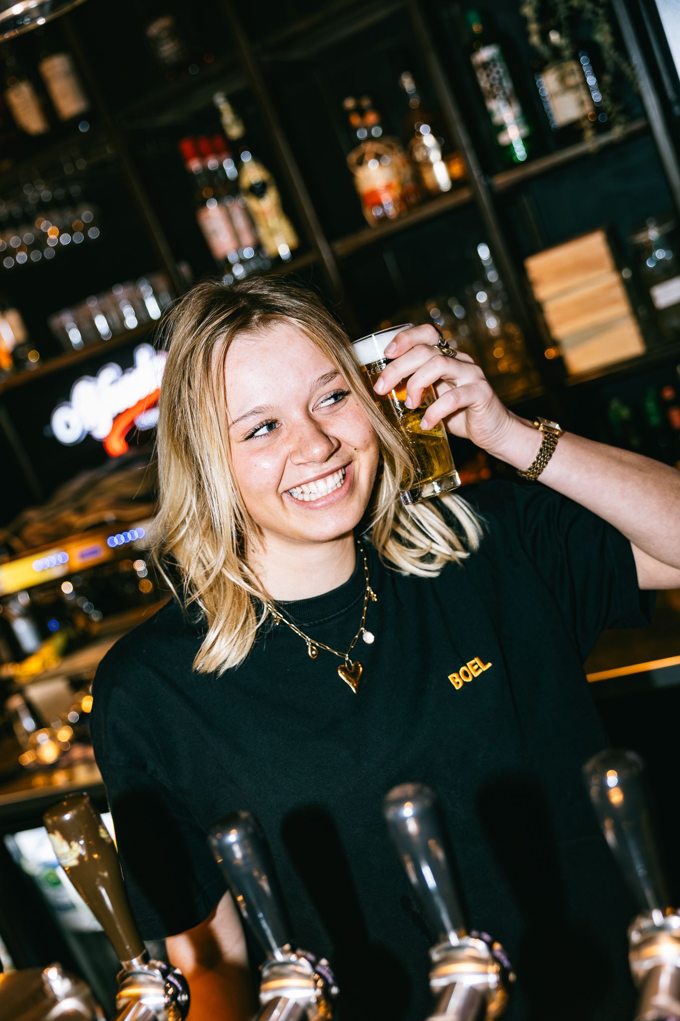 Smiling female bartender in black BOEL shirt holding a pint of beer in front of tap handles at a bar.