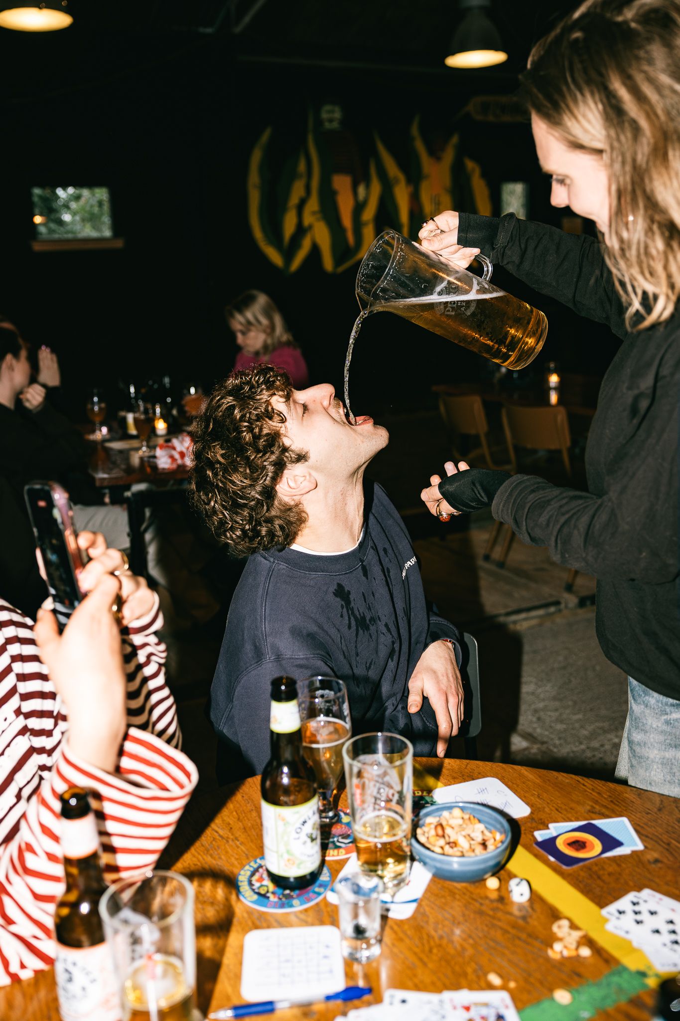 Person having beer poured into their mouth from a pitcher at a bar table with cards and bottles.