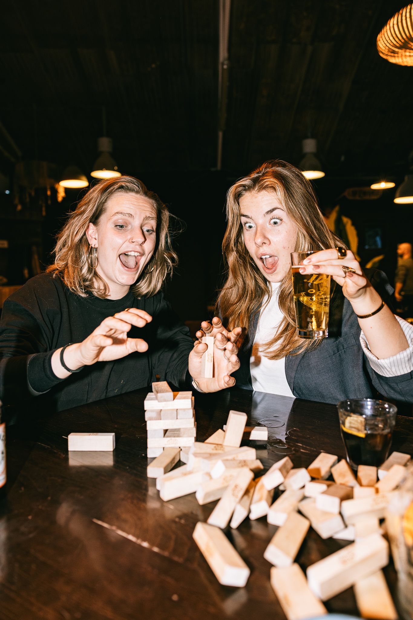 Two women laughing and playing Jenga at a bar, with scattered wooden blocks and a glass of beer on the table.