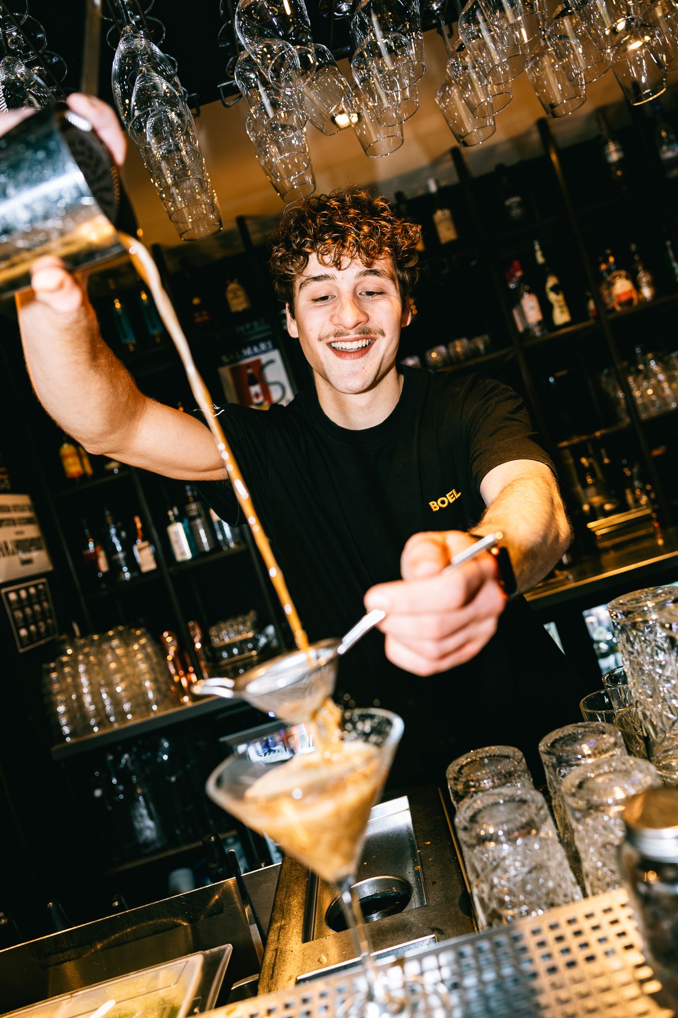 Smiling bartender pouring a cocktail through a strainer into a martini glass at a stylish bar.
