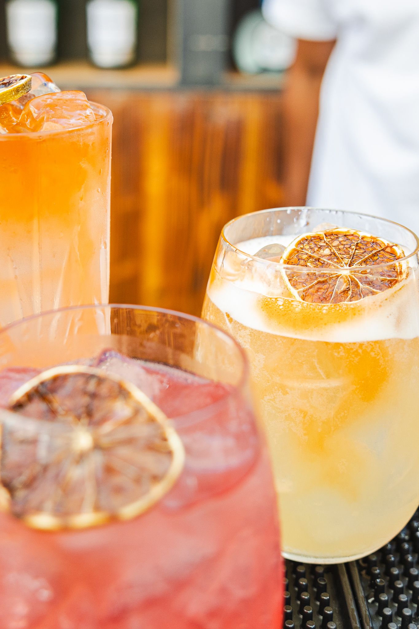 Four colorful citrus cocktails with dehydrated orange garnishes lined up on a black bar mat in warm lighting.