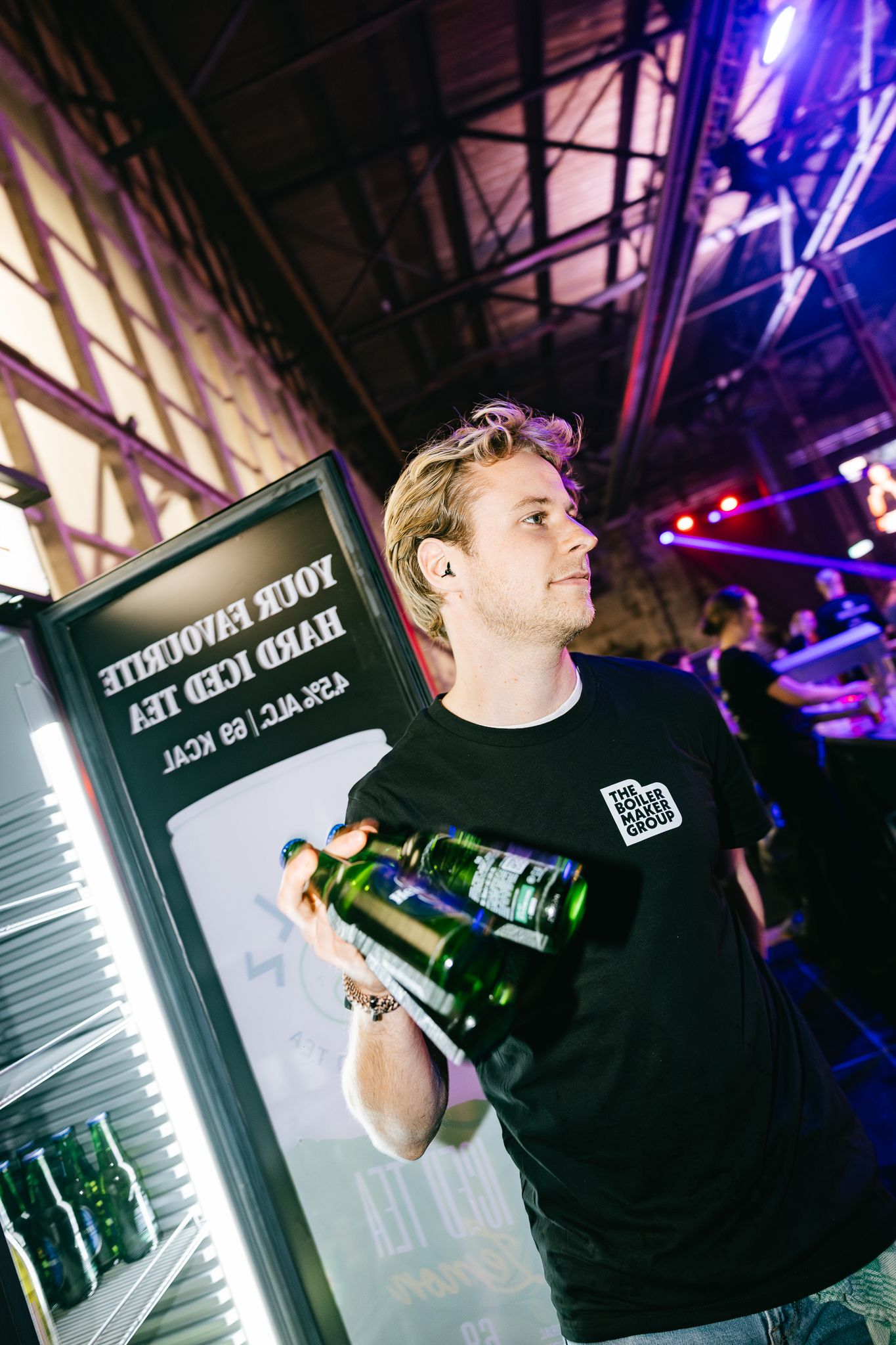 A young man in a black t-shirt holds green bottles near a refrigerator at a vibrant nightclub event.