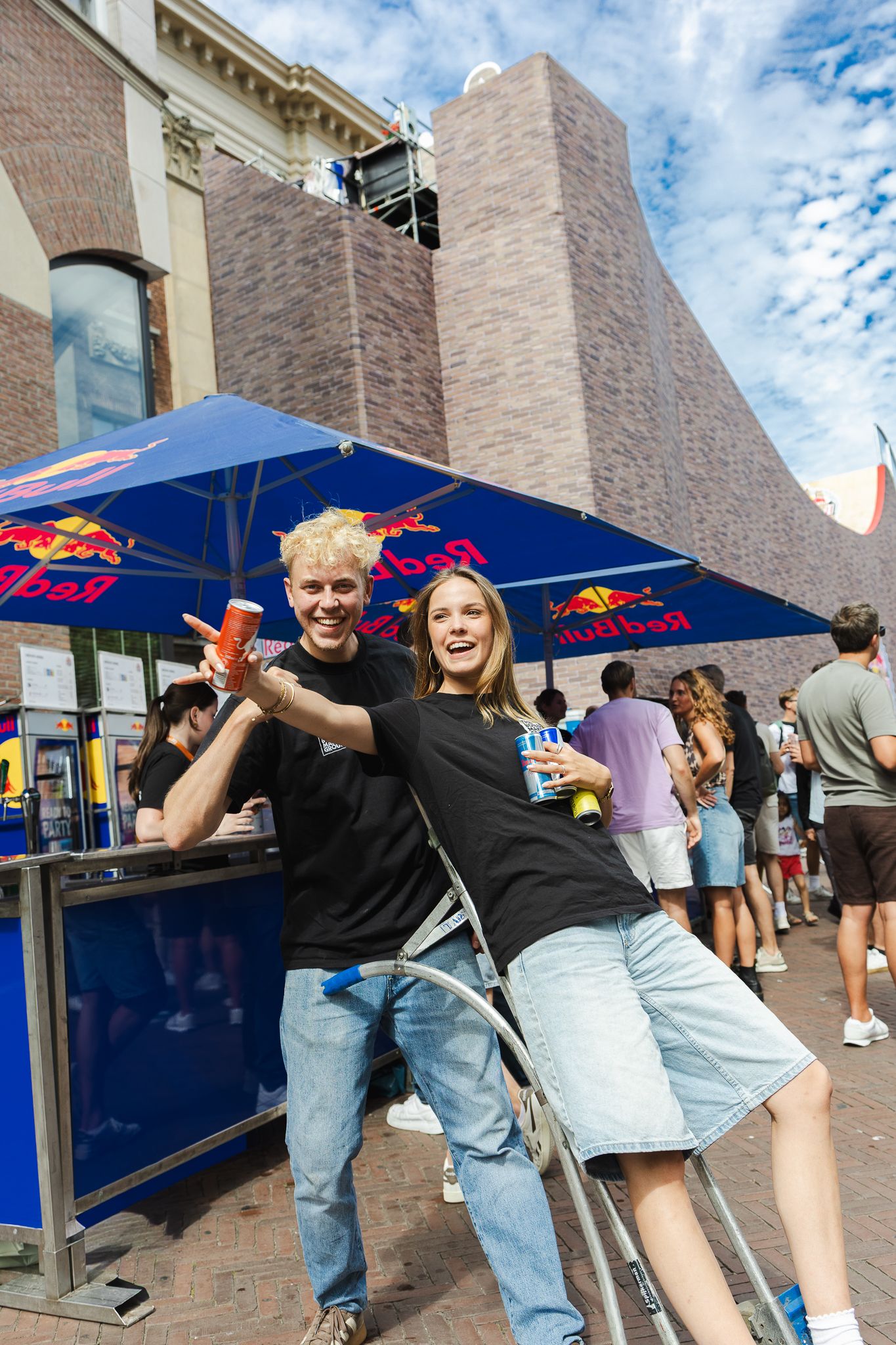 Two smiling friends pose at a Red Bull outdoor event booth, holding cans in black shirts and jeans on a sunny day.