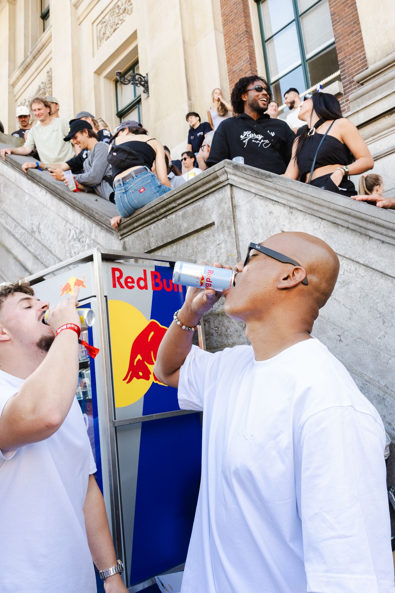 Two men drinking Red Bull energy drinks outdoors at a crowded event near building steps.