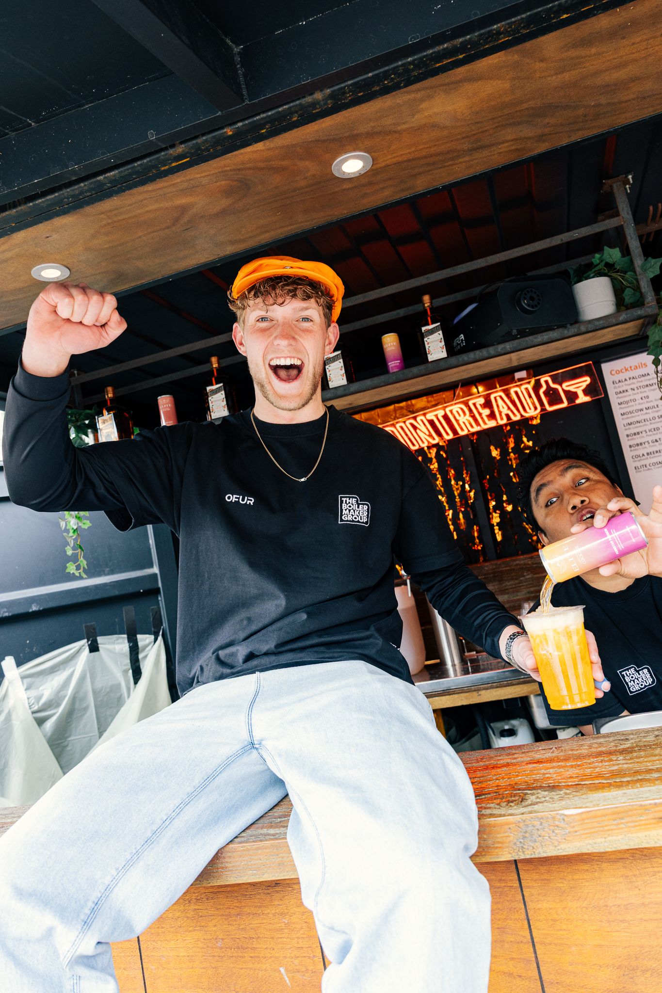 Excited young man in orange cap raises fist at outdoor bar while bartender pours a colorful drink.