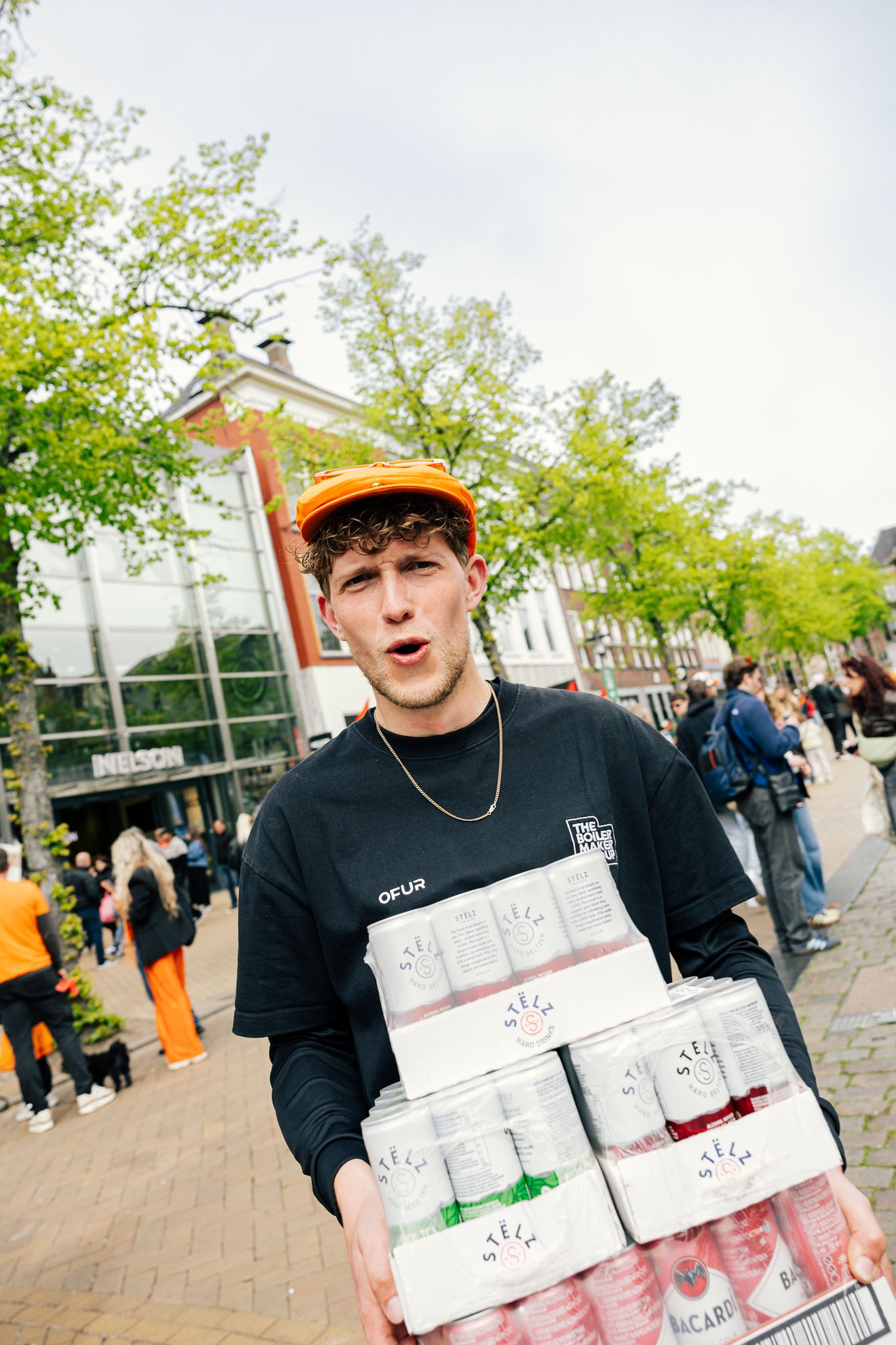 Young man in orange cap carrying cases of Bacardi drinks outdoors at a busy street event.