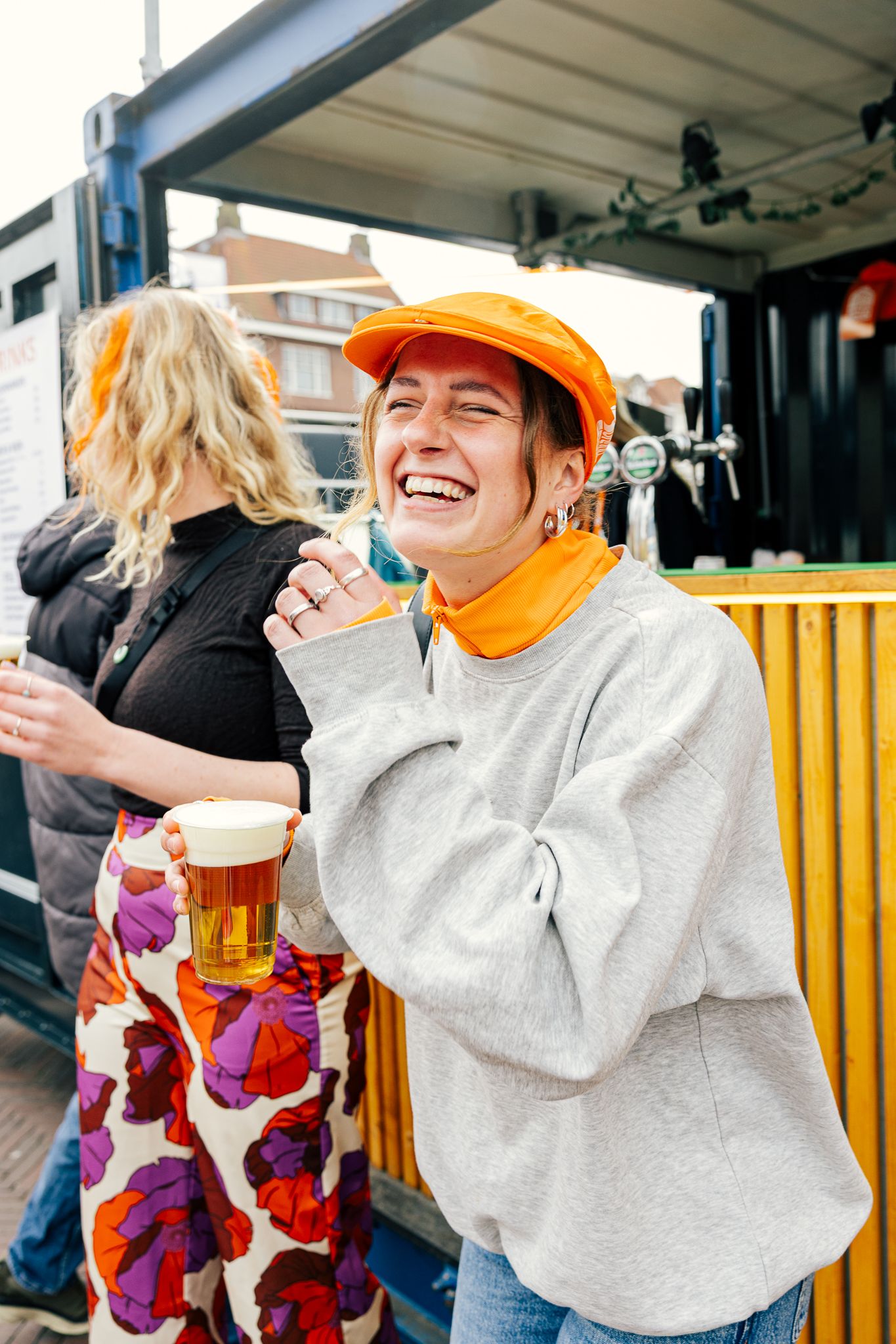 Laughing woman in orange cap holds beer at outdoor festival bar, wearing gray sweatshirt and floral skirt.