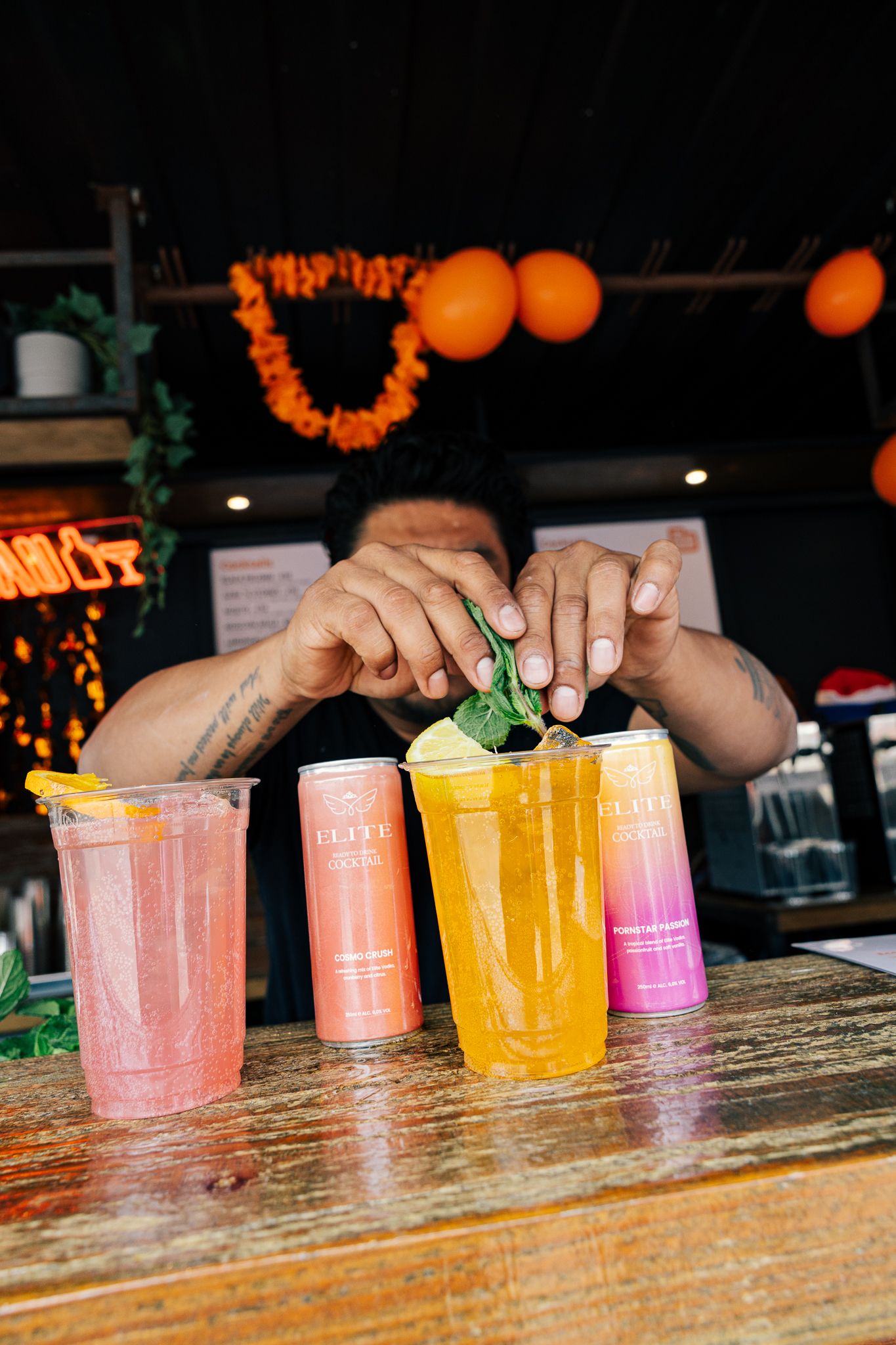 Bartender garnishing colorful cocktails made with RECESS sparkling water cans on a wooden bar with orange decor.