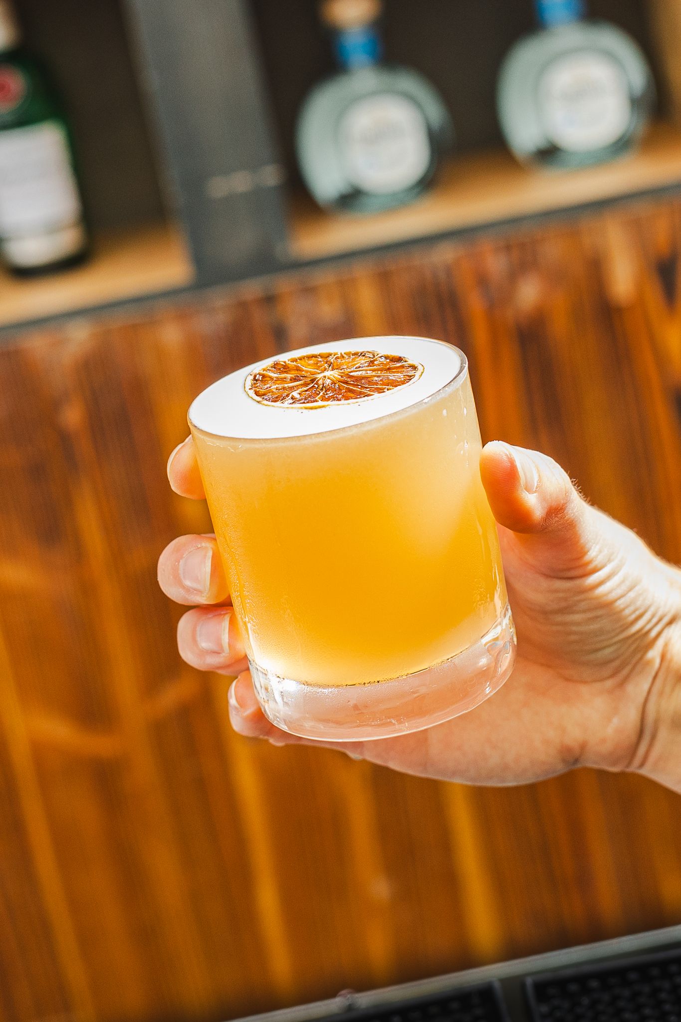 Hand holding a chilled craft cocktail with citrus garnish and frothy top in a rocks glass against wooden bar background.