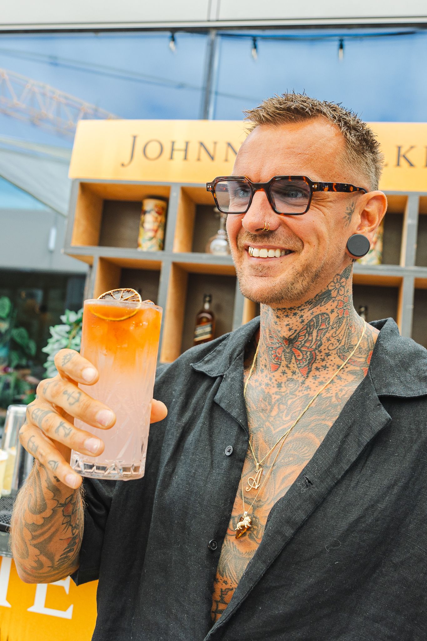 A person with tattoos smiles while enjoying a beer at an outdoor patio.