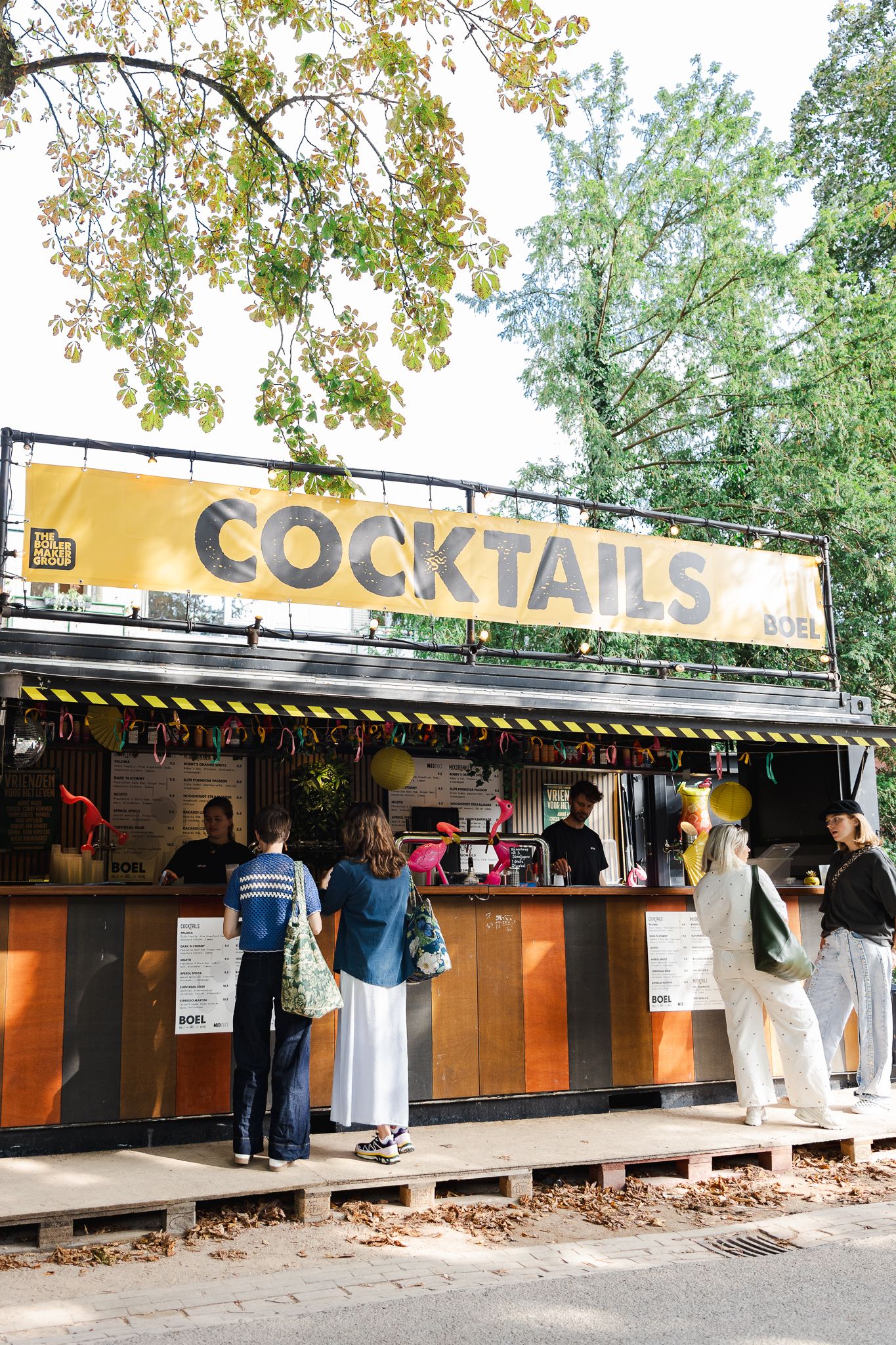 Outdoor cocktail bar stand with yellow signage under trees with customers ordering at the counter.