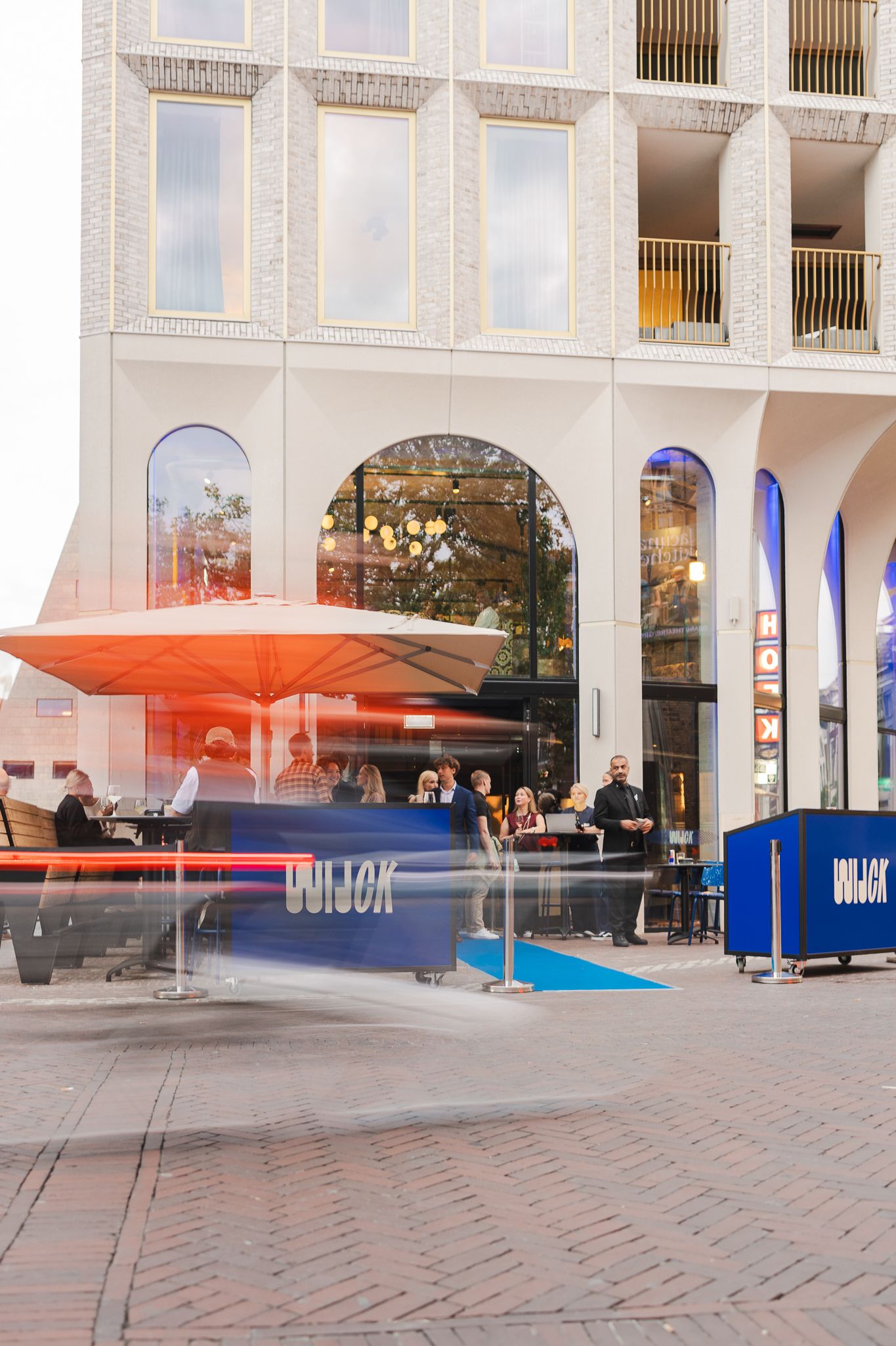 Modern indoor mall interior with orange umbrellas and blue benches arranged in a spacious brick-floored atrium.