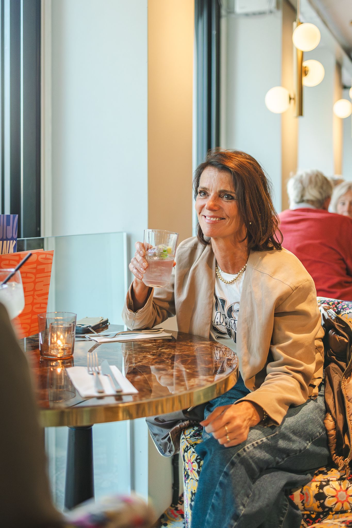 A person in casual attire smiles while sitting at a restaurant table near a window with warm lighting.