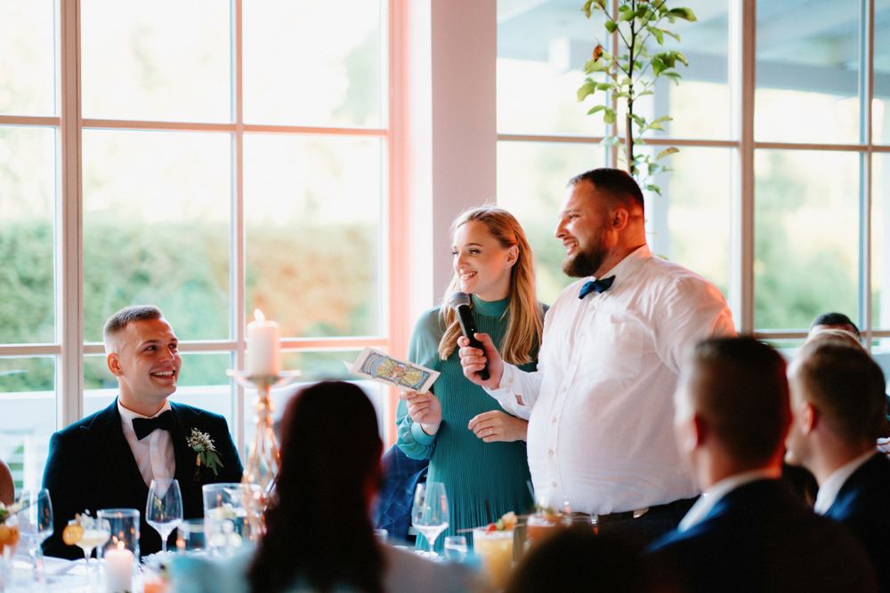 Wedding reception speech with guests seated at tables in natural light.