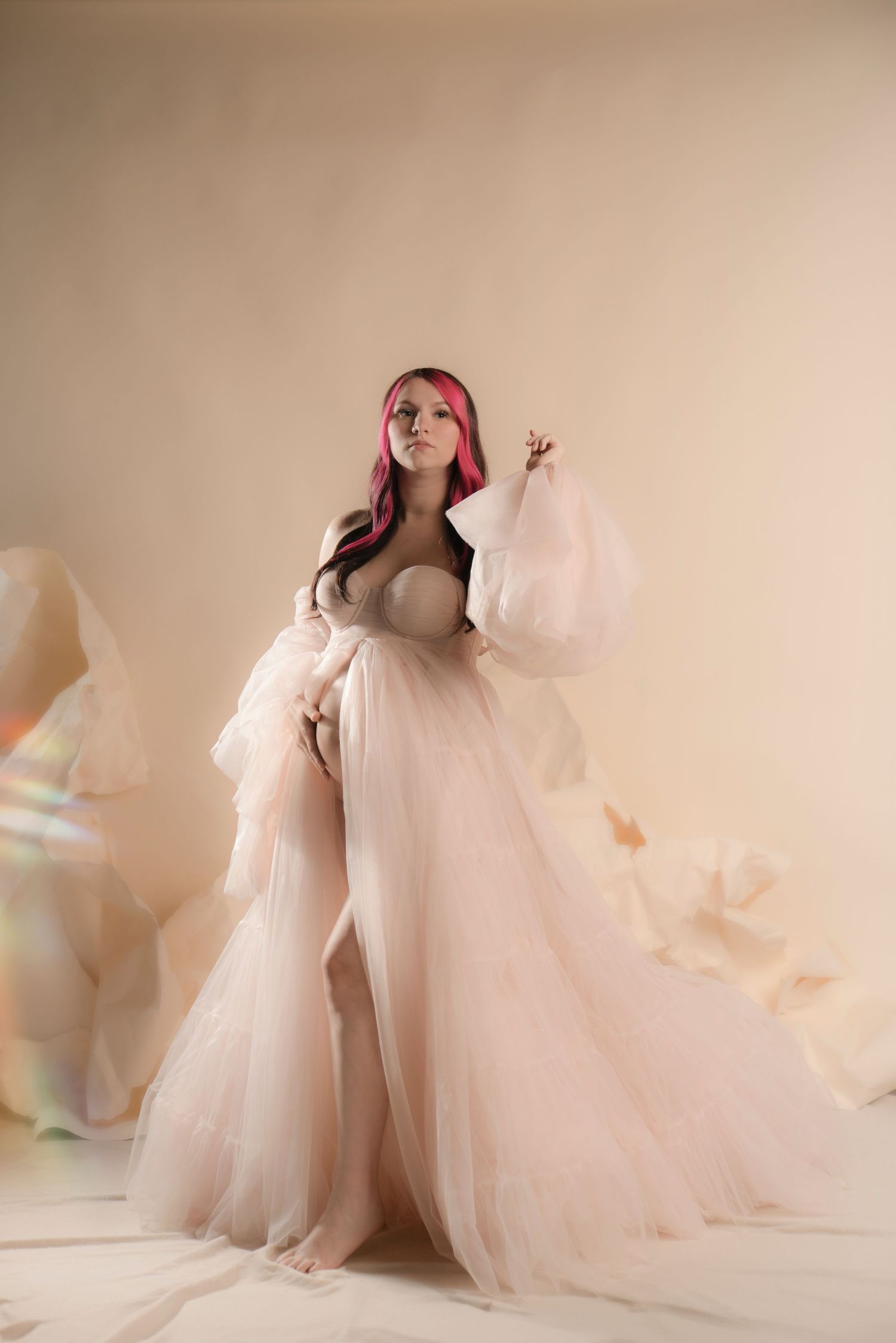 Ethereal maternity portrait featuring flowing sheer blush tulle gown and romantic lighting against neutral backdrop.
