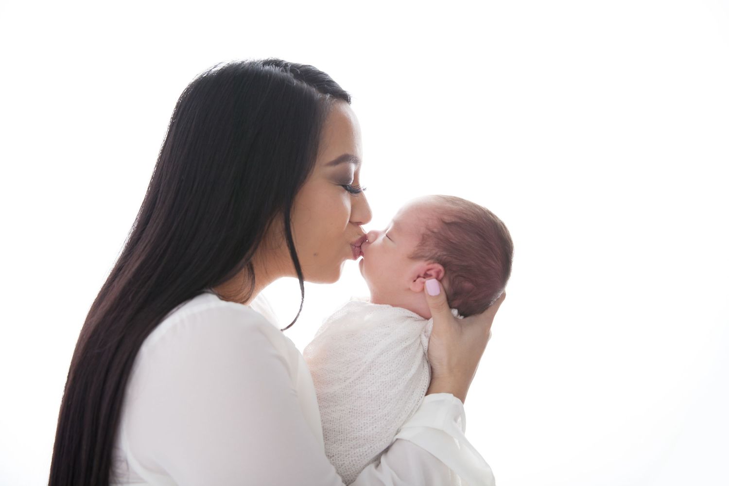 Parent kissing newborn baby wrapped in white against light background.