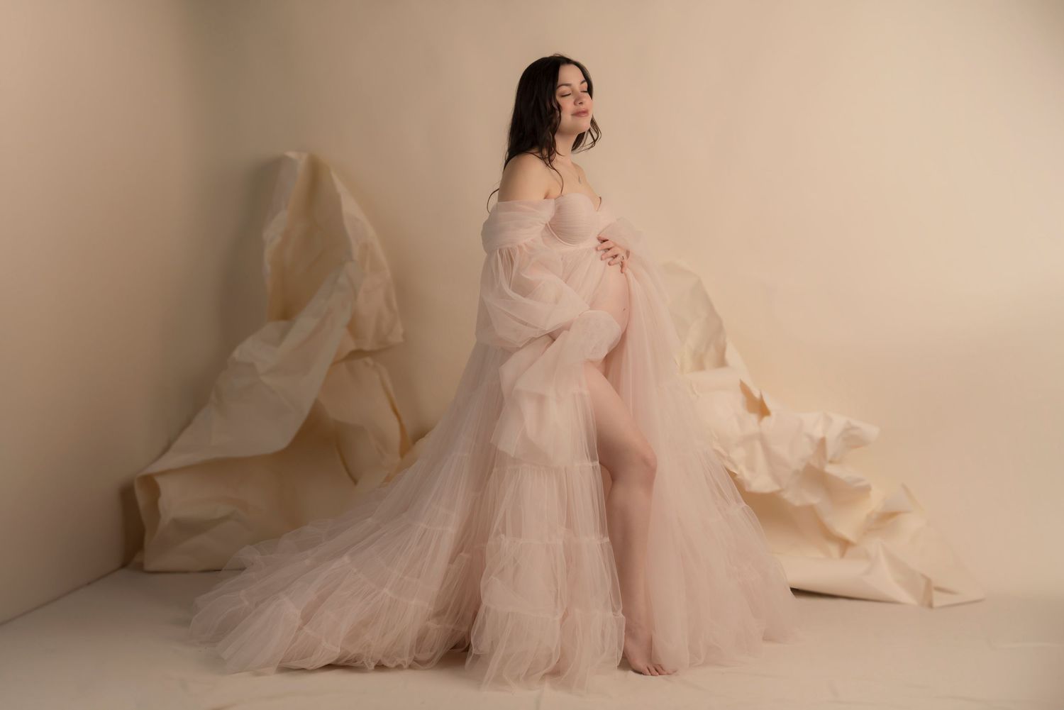 A pregnant woman in a flowing blush pink tulle gown poses for a maternity photoshoot against a beige backdrop. Northeast ohio