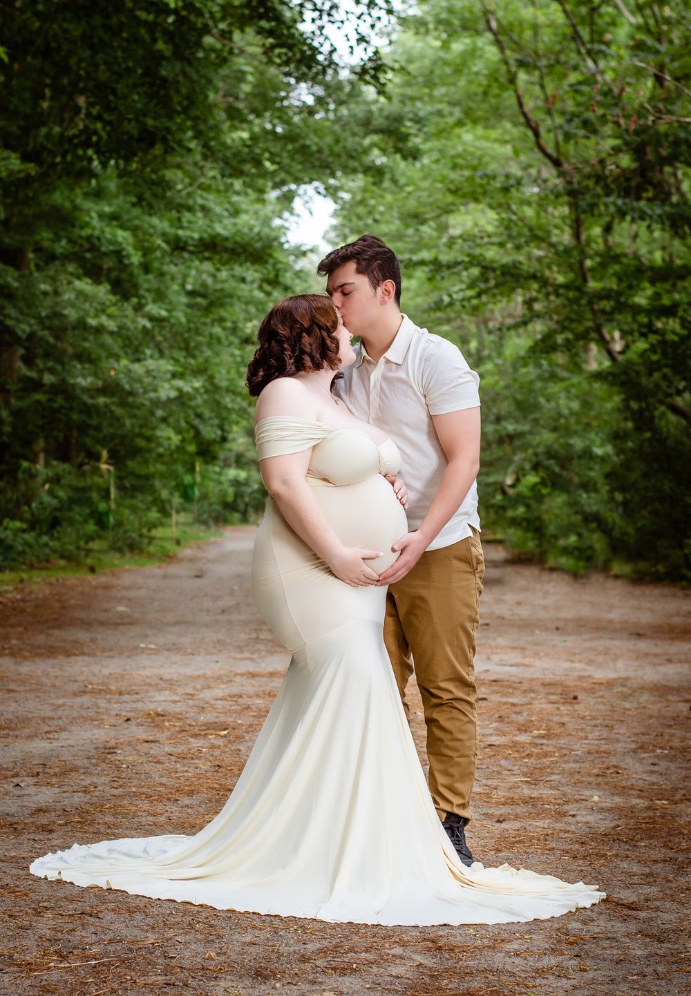 A romantic maternity photo shows a couple embracing on a wooded path, with the pregnant person wearing a flowing white dress.