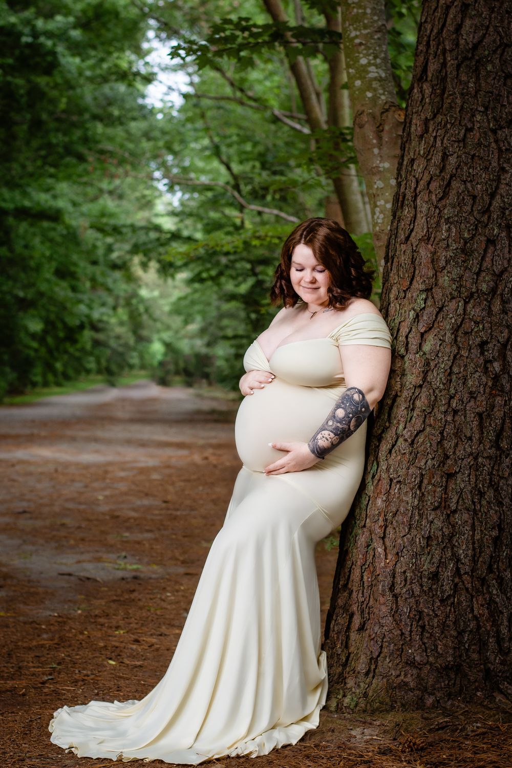 Maternity portrait of an expectant parent in a white dress leaning against a large tree trunk in a forest setting.