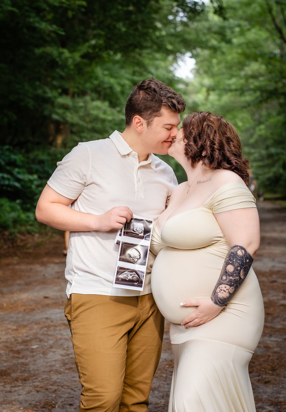 A couple shares a tender moment while holding ultrasound photos during their maternity photoshoot on a forest path.