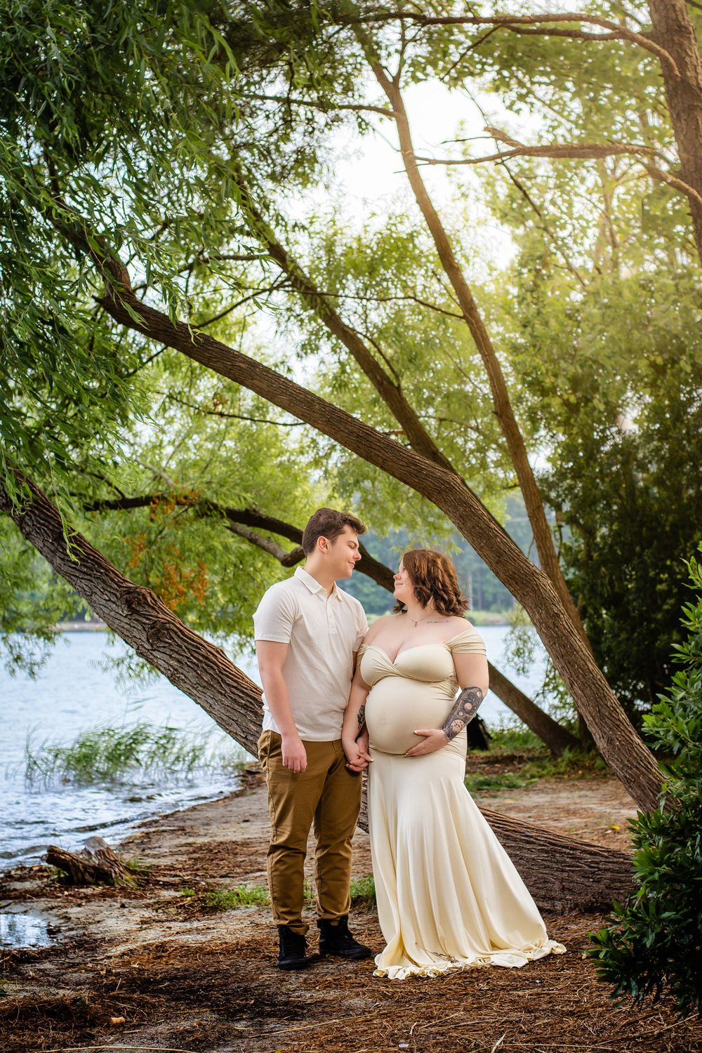 A maternity photoshoot by a lake features a couple in coordinated outfits under leaning trees.