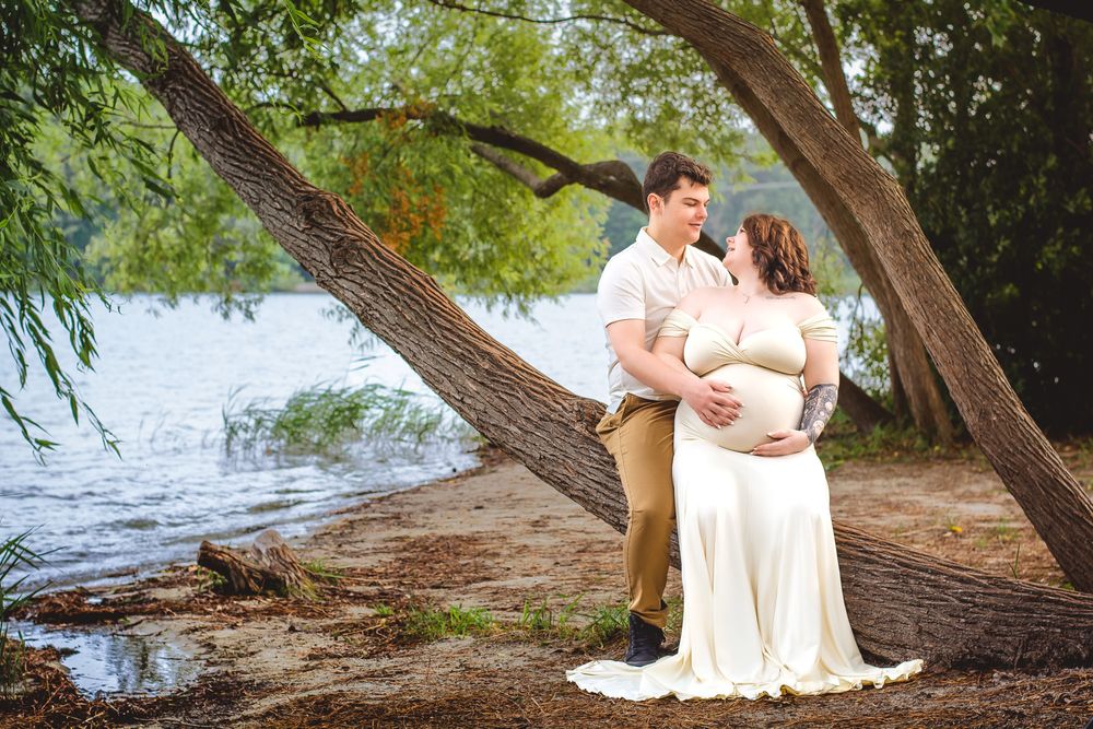 A serene lakeside maternity portrait captures a couple's tender moment under arching trees.