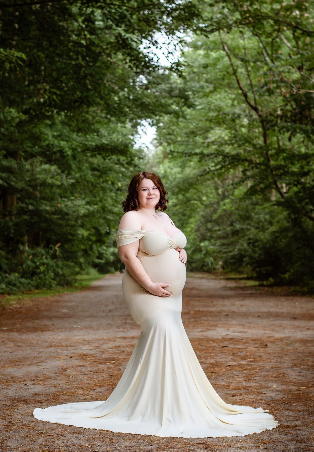 An elegant maternity portrait showcases an expectant parent in a white fitted dress on a forest path.